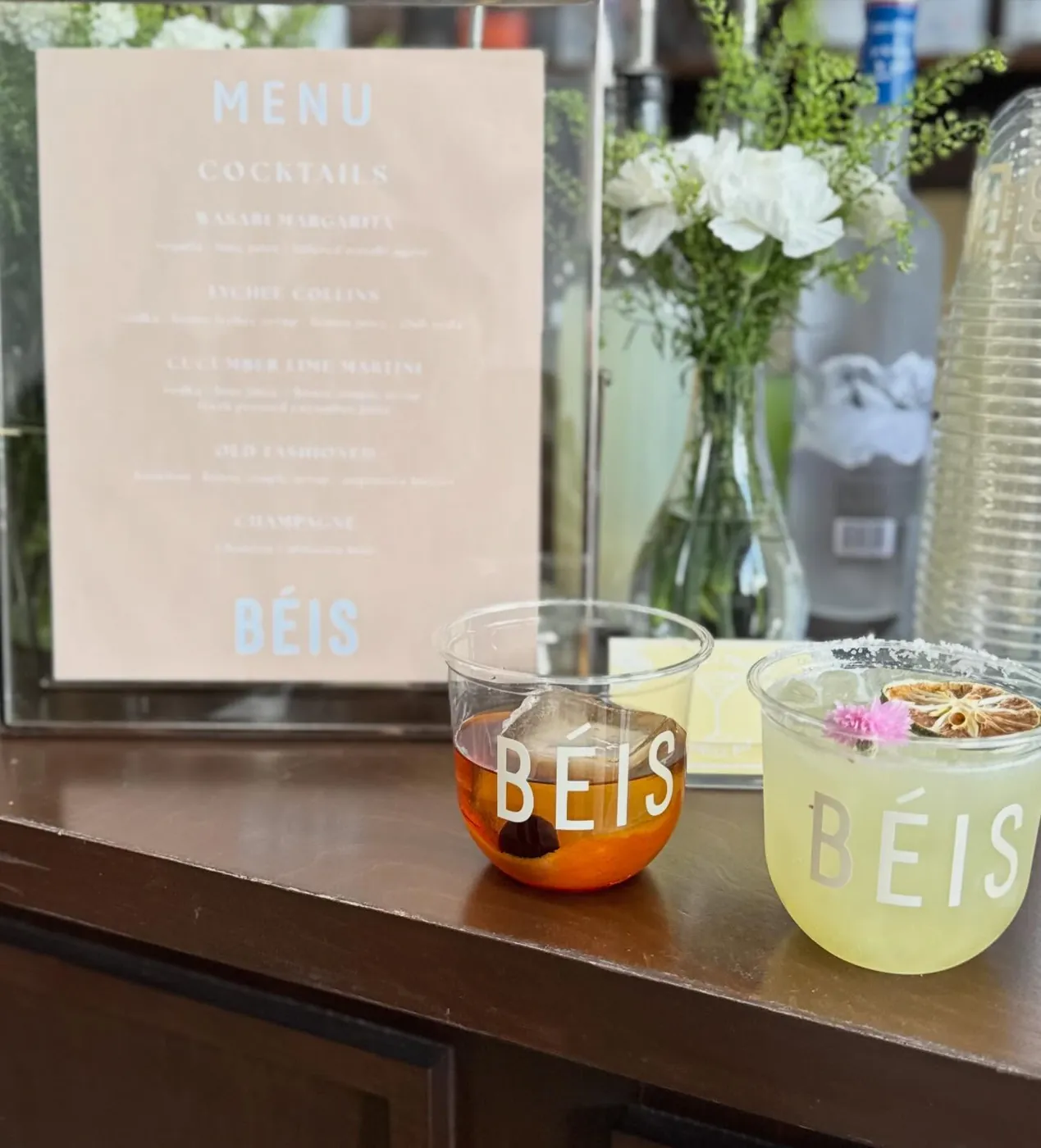 A printed BÉIS bar menu beside branded cocktail cups and white florals