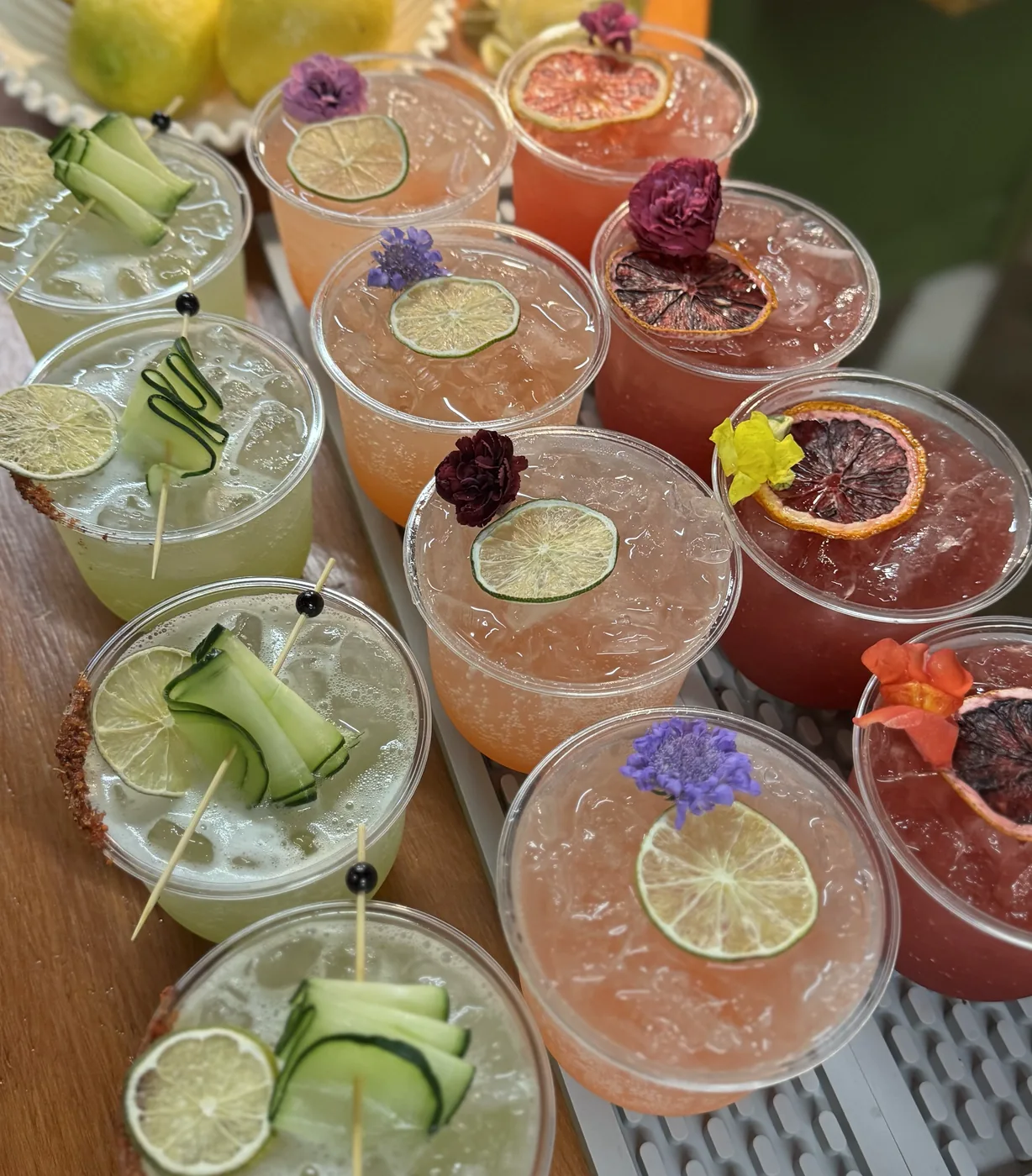 Cocktails lined along a wooden bar beside a bowl of lemons, each garnished with citrus and flowers