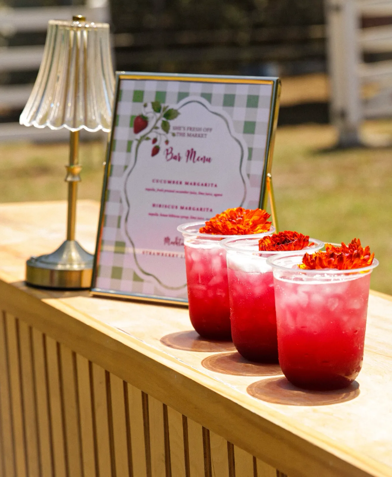 A printed bar menu and brass lamp beside three hibiscus margaritas topped with marigold